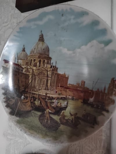 This is a circular decorative plate or platter featuring a painted scene of Venice, Italy. The artwork depicts a canal with several gondolas, some occupied by people. In the background, a large, ornate building with domes and intricate architectural details dominates the skyline, suggestive of a grand church or palace. Buildings with reddish-brown facades line the canal. The sky is painted with clouds, indicating daytime.
The scene captures a bustling waterway, with several gondolas navigating the water. One gondola in the foreground has a figure standing and appearing to row, while others are either moored or carrying passengers. The overall atmosphere suggests a lively, historical European setting. The artwork has a slightly aged, classic aesthetic. There is no visible text on the plate.