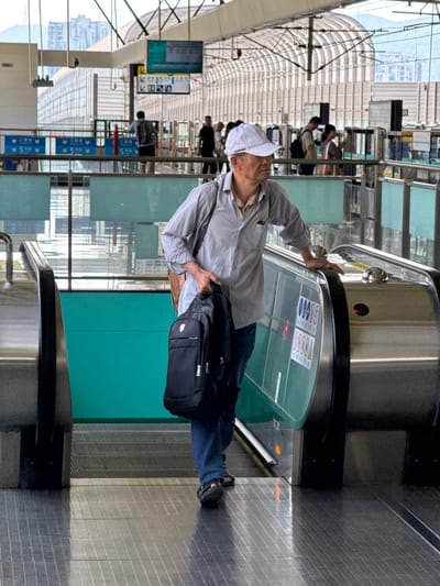 This image, when viewed upright, depicts an elderly person riding an escalator in a brightly lit, modern indoor setting, likely a train station, airport, or large public building.
The main subject is an older individual, presumably male, seen from the waist up. He wears a white baseball cap, a light-colored (possibly light grey or off-white) long-sleeved button-up shirt, and dark blue trousers. He is holding onto the metallic handrail of the escalator with his right hand, and a black shoulder bag or messenger bag hangs on his left side. His expression is neutral, and he appears focused on his ascent.
The escalator itself features metallic steps and a brushed metal handrail. Along the side panels of the escalator, below the handrail, there are transparent green sections. Visible on these panels are white-on-blue pictograms and red-on-white prohibition signs with red circles and diagonal lines, suggesting safety instructions (e.g., prohibiting certain actions like carrying large luggage or children incorrectly). The escalator steps are made of ribbed metal.
In the background, the scene reveals a spacious, contemporary architectural environment with high, possibly arched, ceilings that incorporate large glass panels, allowing ample natural light to fill the space. Through these glass panels in the distance, glimpses of urban buildings and an overcast sky are visible. Numerous other people are present in the background, some walking, others on escalators or moving walkways, though they are less distinct due to depth of field. Several signs are also visible in the background, including a blue sign with white numbers and arrows, such as "5", likely indicating platform numbers or directions within the transportation hub. The bright lighting suggests it is daytime. The overall atmosphere is functional and bustling, typical of a public transport interchange.