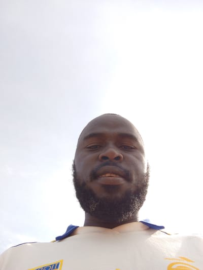 A low-angle selfie photograph shows a man with dark skin, a bald head, and a full black beard with some grey hairs. He is wearing a white shirt with blue trim around the collar. The background is a bright, overcast sky. There is no information in the image to determine the exact location within Mulke Barikin Sayejo, Nigeria.