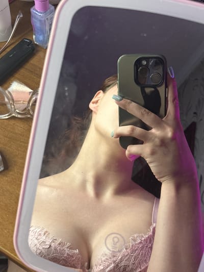 The image is a close-up, low-angle, mirror selfie taken in an indoor setting, likely a bedroom or dressing area. The primary subject is a person, their torso and upper body reflected in a mirror. They are holding a smartphone to take the picture. The person is wearing a light pink lace bralette. A subtle tattoo is visible on their chest. The lighting is a mix of ambient and artificial light, with a pinkish hue casting from the top of the frame, possibly from a light source or a colored filter.
In the foreground, reflected in the mirror, are various objects on a wooden surface. These include a small bottle of what appears to be nail polish or glitter liquid, a black electronic device with some text on it, and a pair of glasses. The background within the mirror reflection is dark and somewhat out of focus, with hints of patterned wallpaper.
The overall mood of the image is intimate and personal, likely intended to be a casual snapshot. There are no clear indicators of time of day or weather conditions. The location context of Bishkek, Kyrgyzstan, is not visually discernible from the image itself.
Visible text on the black object in the reflection appears to read "CHOICE". The text on the bottle is not legible.