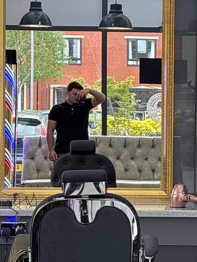 An interior shot shows a barber's mirror reflecting a customer in a black shirt, seated on a tufted seat. The reflection shows an exterior view of a street with a red brick building, trees, and cars, including a white vehicle with visible license plates. The mirror's frame has an ornate gold finish and is decorated with a barber pole pattern along the top. In the foreground, part of a barber's chair is visible, along with a hair dryer. A storefront with the name "CRINGLEFORD" is seen in the reflection. Based on this storefront, the location is likely Cringleford in Norwich, United Kingdom.
