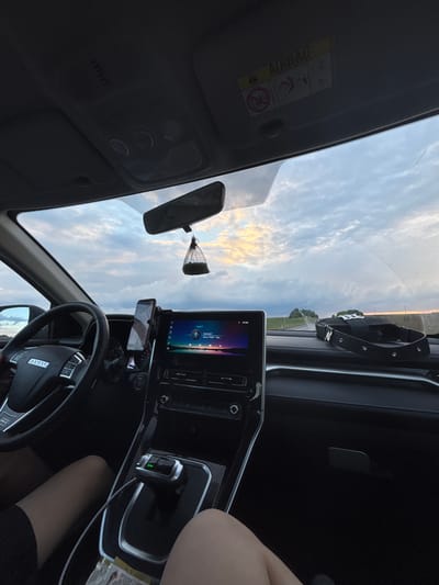 The image shows the interior of a car, looking out through the windshield at a road and a cloudy sky. The dashboard of the car is visible, including the steering wheel, a large infotainment screen, and a gear shift. A phone is mounted on the dashboard. A black bag with a strap is placed on the dashboard. The car appears to be in motion, as indicated by the perspective and the road ahead. There are no visible indications that this image was taken in the city of Krasnoyarsk, Russia.