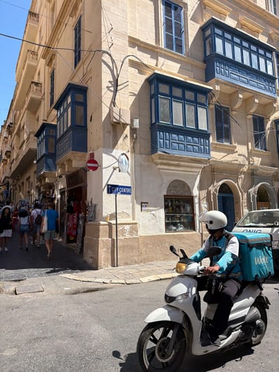 This vertical, full-frame shot captures a street scene in Valletta, Malta, under bright daylight, likely mid-morning or early afternoon given the strong shadows. The image is dominated by the architectural style of the city, with traditional Maltese balconies featuring ornate blue wooden frames and glass panes, set against cream-colored stone buildings.
In the foreground, a person wearing a white helmet and a blue and white uniform is seated on a white scooter. They have a large, turquoise delivery bag with the "Wolt" logo emblazoned on its side attached to the back of the scooter. The rider's posture suggests they are either waiting or have just stopped.
The street itself is paved with dark asphalt, and a sidewalk is visible with several people walking. Shops line one side of the street, with displays of souvenirs and other merchandise visible through their windows. A blue street sign with white lettering indicates "CITY CENTRE" and an arrow pointing forward. A red circular "no entry" sign is also visible on a pole.
The architecture is a prominent feature, with detailed stonework, arched doorways, and multiple floors of windows. Electrical wires are strung across the buildings. The overall impression is a bustling urban environment with a strong sense of place, indicative of a historic European city. The weather appears to be sunny and warm.