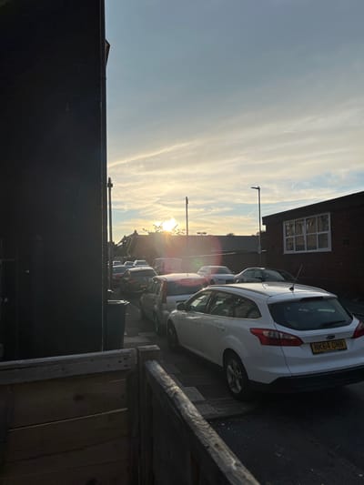 The image shows a street scene in Stoke-on-Trent, United Kingdom, during sunset. A dark building on the left side of the frame casts a shadow over a portion of the scene. In the background, several cars are parked along the street, and houses with pitched roofs are visible. The sky is illuminated by the setting sun, creating a warm glow with streaks of clouds. A streetlamp stands in the middle ground. In the foreground, a wooden fence is partially visible. The license plate of the white car in the foreground reads "NK64 ONN".
