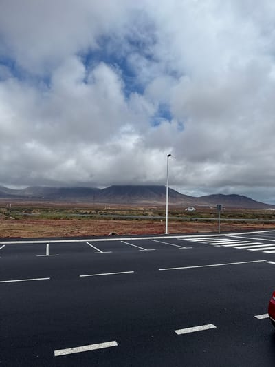 An outdoor scene in Yaiza, Spain, features a vast expanse of dark asphalt in the foreground, likely a parking area or road. This surface is distinctly marked with numerous white lines, indicating parking spaces and a prominent pedestrian crossing or zebra pattern towards the left. A small portion of a red vehicle is visible at the very top right edge of the asphalt.

In the midground, a tall, slender white pole stands vertically, with a small, dark bird perched atop it. Beyond the asphalt and extending into the mid-distance, the terrain is covered in sparse, low-lying vegetation with reddish-brown and green hues, characteristic of an arid or semi-arid environment. A distant white car is visible on a road that traverses this landscape.

The background is dominated by a range of dark, undulating mountains that stretch across the horizon. The sky above is largely overcast with white and grey clouds, though patches of clear blue sky are visible through breaks in the cloud cover. The lighting suggests it is daytime. No discernible text is visible in the image. The scene captures a blend of natural ruggedness and modern infrastructure under a cloudy sky.