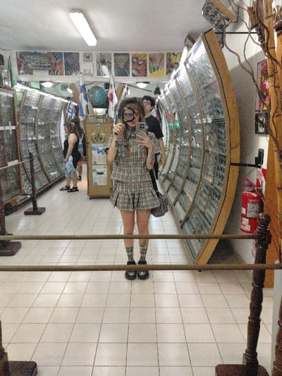 A person with a magnifying glass stands in a hallway. They wear a matching plaid skirt and top with matching socks. Other people are walking down the hallway, which is lined with glass cases. There are also some flags and other decor in the hallway. A mirror cuts through the middle of the image.