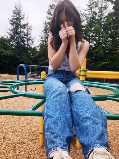 A young female is depicted seated on a green and yellow metal playground structure. She has long, dark brown hair, partially obscuring her face, and is dressed in a white sleeveless top with a light blue floral pattern, distressed blue jeans with tears at both knees, and light-colored athletic shoes. Her hands are clasped together, thumbs extended upwards, positioned in front of her partially obscured face; her eyes appear closed or squinting. The ground beneath the structure is covered with light brown wood chips or mulch. The background consists of numerous tall evergreen trees and an overcast sky. Additional playground equipment, including a blue metal structure and a distant swing set, is partially visible. The exact location within Keskanterak, Uzbekistan, cannot be determined from the visual information provided.