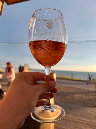 The image depicts a close-up, first-person perspective of a hand holding a glass of sparkling rosé wine outdoors during sunset in Vyshesteblievskaya, Russia. The clear glass is filled with a pale pink, bubbly liquid, with the word "FANAGORIA" and "NOBLESSE OBLIGE" clearly visible on its side, alongside a distinct, patterned emblem. The hand holding the glass has light pink, speckled nail polish.

In the background, a bright, clear sky transitions from light blue overhead to a warm, soft orange hue near the horizon, indicative of late afternoon or early evening. A blurred fence or railing separates the sky from what appears to be a body of water or distant land. Vague, out-of-focus figures of people are visible in the far distance, suggesting a public area like a waterfront promenade, pier, or outdoor terrace. Parts of a wooden surface, possibly a table or deck, are visible in the foreground and to the right. The scene conveys a relaxed, leisurely atmosphere.
