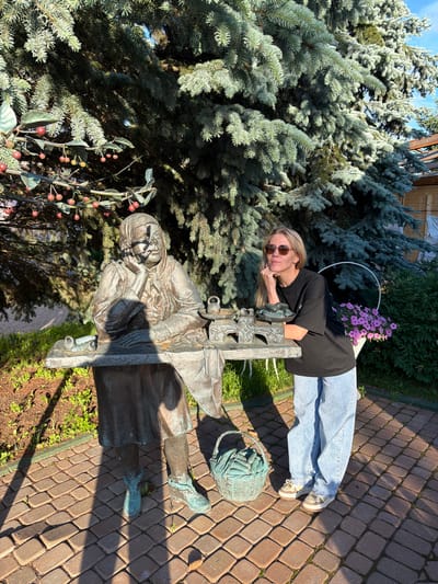 A young woman with blonde hair and sunglasses is leaning on a bronze statue of an elderly woman. The woman is wearing a black t-shirt, light blue jeans, and sneakers. She is looking at the camera with a slight smile. The statue is of an elderly woman wearing a headscarf and a long dress, with her hands resting on her lap. The statue is located outdoors, next to a large blue spruce tree. In the background, there are pink flowers in a white pot, a wooden fence, and a paved walkway. The scene appears to be set on a sunny day, likely in the late afternoon due to the long shadows. The location is Suzdal, Russia. There is no visible text in the image.