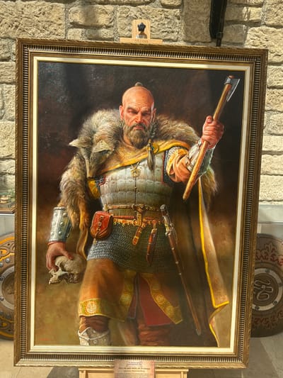 The image displays a framed oil painting of a fearsome warrior standing in front of a stone wall. The warrior, with a shaved head and beard, wears a fur-lined cloak over intricate armor and chainmail. He holds an axe in his right hand and a human skull in his left. The painting is set up on a wooden easel, with a small placard at its base. In the background, two shield-like objects with ornate carvings are visible on either side of the painting. The overall scene appears to be an exhibition or display, likely in a museum or gallery, as suggested by the placard and the context of the stone wall. The placard at the bottom of the painting is in Bulgarian and appears to provide information about the artwork and its artist, Miroslav Yotov, possibly in relation to the city of Neofit Rilski, Bulgaria.