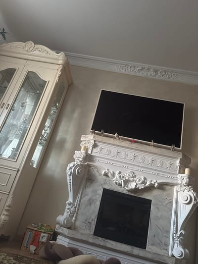 The scene shows an interior with elaborate white decorative molding, including ceiling trim, a mantlepiece featuring a mounted flat-screen television above what appears to be a faux fireplace insert. A white cabinet with glass doors is also visible. A child's toy is on the floor near the right side of the image. The location cannot be determined solely from the image. There is no evidence of the image being taken in Bakhchysarai, Ukraine.