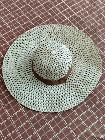 A straw sun hat with a brown ribbon band rests on a brown and white checkered tablecloth. The hat has a wide brim and a round crown. The background is a close-up of the tablecloth, showing the diamond pattern. There are no people or animals visible in the image. The image is taken from a high angle, looking down at the hat. The lighting is even and natural. The location is not specified in the image.