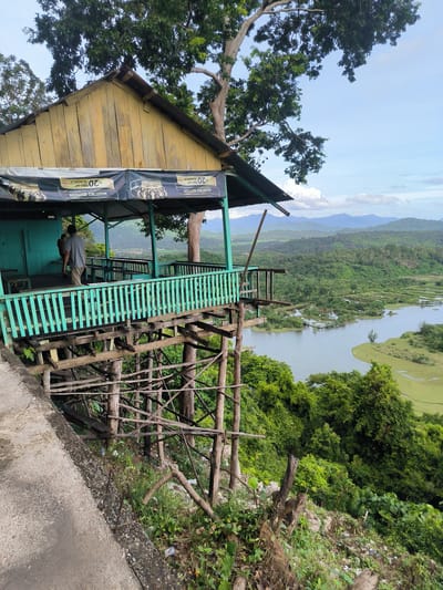 This is a landscape image taken from a high vantage point, overlooking a lush, green valley with a river or lake winding through it. In the foreground, a weathered, yellow wooden building with a green railing stands precariously on stilts made of rough-hewn timber. A man in a gray shirt and beige pants stands on the wooden deck of the building, facing away from the camera. The building appears to be a simple structure, possibly a cafe or resting spot, with a tarp banner overhead advertising "MAESTRO EDITION" and "20 RIBU/BUNGKUS". The surrounding vegetation is dense and green, with tall trees framing the scene. The background shows rolling hills and mountains fading into the distance under a partly cloudy sky. The overall impression is one of a rustic, somewhat neglected establishment offering a panoramic view of the natural landscape. The location is identified as Meudheun, Indonesia.