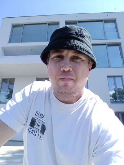 In Doksy, Czechia, a person wearing a black bucket hat and a white t-shirt with a graphic of a boombox is captured in a selfie. The person is positioned in the foreground, with a modern white building featuring large windows in the background. The sky is clear and blue. The image is taken outdoors on a sunny day.