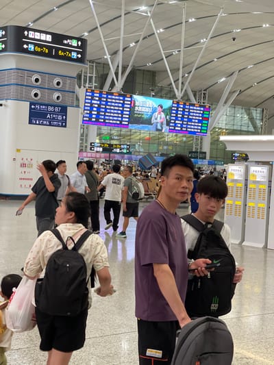 The image captures a scene inside what appears to be a busy transportation hub, possibly an airport or train station, given the departure boards and signage. The primary subjects are several individuals standing or walking, some with luggage, suggesting they are travelers. Two young men in the foreground are prominently featured, looking towards the camera. One is wearing a purple t-shirt, and the other a white t-shirt with text. Behind them, other travelers are visible, including people with backpacks and shopping bags.
The setting is indoors, characterized by a modern architectural style with a high ceiling and bright, artificial lighting. Multiple large digital display boards are visible, showing flight or train information, with columns listing departure times, destinations, and other relevant details. One board displays Chinese characters, including "越往山里去 水质就越好" which translates to "The further into the mountains, the better the water quality." Another sign reads "深圳欢迎你," meaning "Shenzhen Welcomes You." Charging stations for mobile phones are also visible, labeled "手机充电站" (mobile phone charging station).
The overall atmosphere appears to be one of transit and waiting. The individuals in the foreground seem to be posing for a photo, with the younger man in the white shirt smiling faintly. Other people in the background are more focused on their journeys, walking or standing in queues. The lighting suggests it could be daytime or evening, but the artificial illumination makes it difficult to determine the exact time of day. The presence of multiple departure boards and the general bustle indicate a public space with high foot traffic.