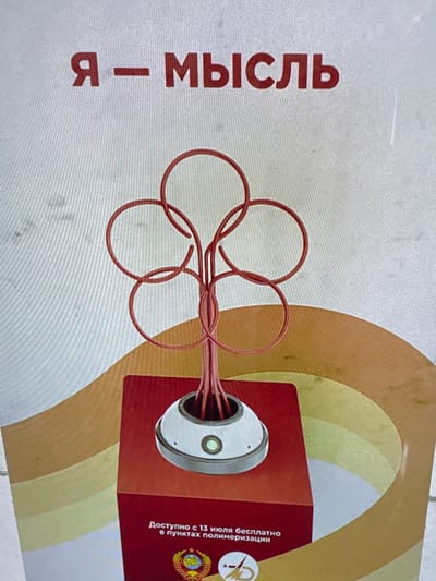The image displays a digital graphic, likely a poster or advertisement, featuring Cyrillic text and an abstract design. The primary elements include large, vertical red text on the left, reading "Я – МЫСЛЬ" (I – THOUGHT), set against a textured light grey background.

To the right, a complex network of red wires forms several loops, resembling a stylized brain or neural connections. These wires appear to emanate from a metallic, circular component embedded within a reddish-brown square box. The background behind this box transitions into soft, curved bands of light brown and yellow-green.

Smaller vertical text on the right side of the red box, also in red, states: "Доступно с 13 июля бесплатно в пунктах полимеризации" (Available from July 13 for free at polymerization points). Below this text, a small emblem is visible, which closely resembles the State Emblem of the Republic of Belarus, featuring a globe, sun, wheat stalks, and a red star.

No people are present in the image. The graphic conveys a conceptual theme, possibly related to thought, technology, or an art installation, with specific details about its availability. The Russian language and the Belarusian emblem suggest a cultural or informational context originating from that region, potentially displayed in Katwijk, Netherlands. The time of day, weather, or emotions are not discernible from this static graphic.
