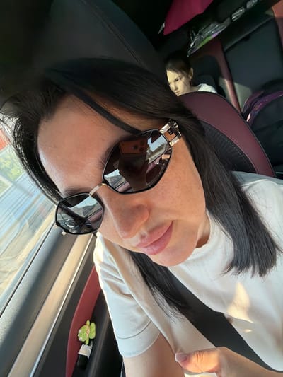 A woman in sunglasses and a light-colored t-shirt is in the driver's seat of a car, looking down. Her phone, with a case, is visible in the reflection of her sunglasses. A child with dark hair and a white shirt is in the back seat. The car's interior is dark with red accents. A plush toy is clipped to the car's interior panel. The scene appears to be on a road, as suggested by the reflection in the sunglasses. The image does not provide any specific information about the city of Dol "Luch", Russia.