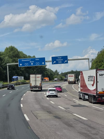The image shows a multi-lane highway with traffic. Overhead signs indicate directions to Düsseldorf, Bonn, and Köln (Cologne), and Trier and Koblenz. Several trucks and cars are visible on the road. The environment is characterized by a blue sky with white clouds and dense green trees lining the sides of the highway. The weather appears to be clear and sunny. This scene is likely a portion of the A3 autobahn near Dernbach (Westerwald), Germany, a known junction for these routes.