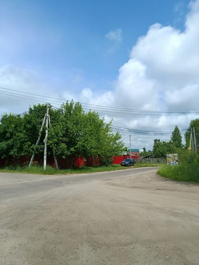 The image shows a view of a dirt road intersection in a rural area, possibly the city of Тополиный, Russia. A dark blue SUV is parked on the left side of the road. A bright red fence runs along the left side of the road, behind which are dense green trees. On the right side of the road, there is tall green grass and a wooden fence. Several utility poles and power lines are visible throughout the scene. The sky is blue with scattered white clouds. The overall scene depicts a quiet, possibly residential, area.
