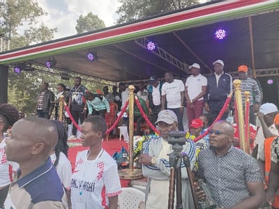 A crowd of people are gathered in front of a raised stage. On the stage, several men stand, including one in a "PRADA" t-shirt and another wearing a baseball cap. The stage is decorated with lighting and a banner with red, white, and green stripes. A red velvet rope barrier is in place. In the foreground, a man with sunglasses stands holding a camera and a man stands next to a tripod.