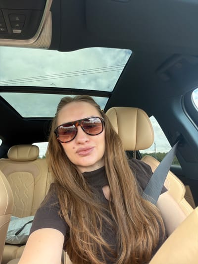 This is a selfie taken by a woman inside a car. The woman is wearing sunglasses with tortoiseshell frames and a dark brown top. Her long, brown hair is spread across her shoulders and chest. The interior of the car features beige leather seats and a dark headliner with a sunroof. A gray seatbelt is visible across her chest. The scene appears to be a daytime shot, with bright, overcast conditions visible through the car windows. Outside the car, power lines and trees can be seen, suggesting a semi-urban or rural setting. The woman's expression is neutral, and the overall mood is casual. No visible text is present in the image.