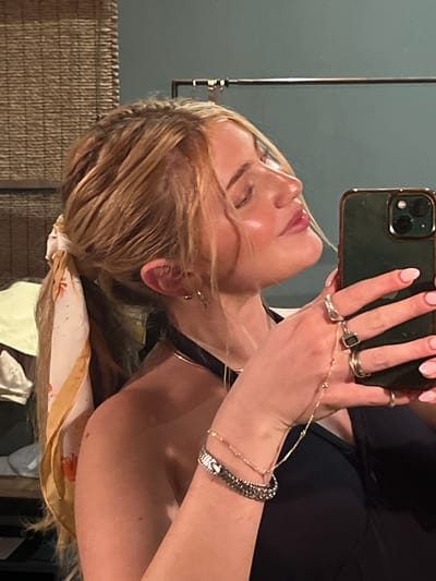 A selfie taken in an indoor environment. A woman with blonde hair and fair skin holds a phone, her hand adorned with multiple rings and bracelets. She wears a black top and a patterned scarf is tied in her hair. The background includes a textured wall. The location is unknown, and there is no information indicating that the image was taken in Livingstone, Zambia.