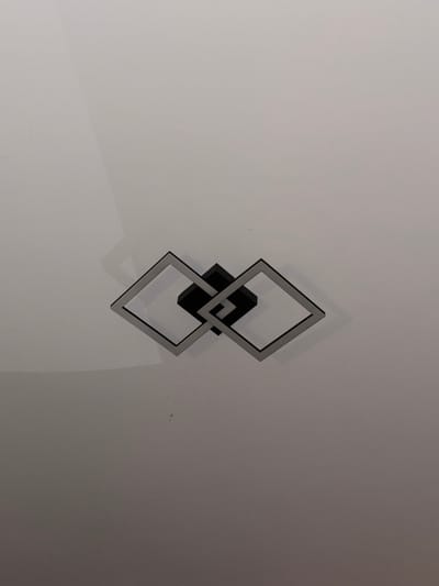 A modern, wall-mounted decorative object is prominently featured on a plain, light-colored wall. The object comprises two interconnected, square-shaped frames. The inner frame is black, seemingly serving as the base mounted to the wall. Overlapping and positioned slightly forward of the black frame is a sleek, light grey or silver frame. This outer frame appears to be illuminated from within, possibly by an integrated LED strip, casting a soft glow. The interplay of the two frames creates a dynamic, three-dimensional effect, with subtle shadows visible on the white wall behind, hinting at the object's depth and an internal light source. The wall is unadorned, suggesting a clean, minimalist indoor setting. The photograph itself is slightly angled. There are no people, visible text, or specific activities depicted. The scene conveys a contemporary aesthetic within an interior space, likely in Levski, Bulgaria.