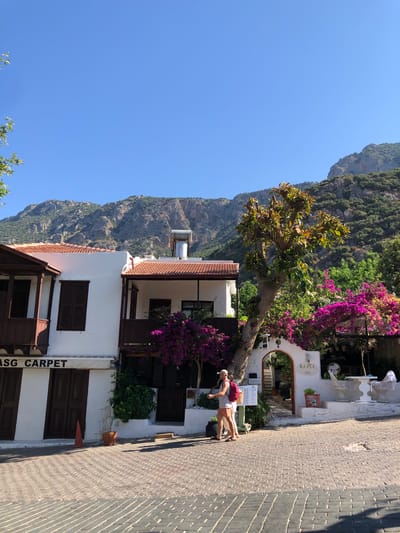 In Kaş, Turkey, a cobblestone street is lined with traditional white buildings adorned with terracotta roofs. A man wearing a beige cap, a white tank top, and orange and white striped shorts walks on the street, carrying a red backpack. He is captured mid-stride, looking towards a narrow alleyway framed by a stone archway and lush pink bougainvillea flowers. The alley leads to a small courtyard with white outdoor furniture and a sign that reads "BAHCE". A large tree with vibrant green leaves stands beside the archway. To the left, a white building features dark wooden shutters and a balcony with dark wooden railings. A sign above the entrance reads "ASG CARPET". The scene is set against a backdrop of rugged, tree-covered mountains under a clear blue sky.