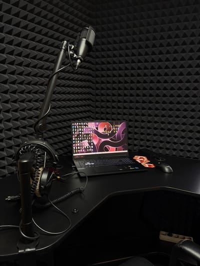 The image shows a recording setup within a soundproof room, likely a home studio. The walls are covered with black acoustic foam panels, creating a textured, egg-crate appearance. In the foreground, a black microphone is mounted on an adjustable boom arm, positioned in front of the foam. The microphone is angled slightly upwards.

The boom arm is attached to a larger, dark-colored structure, possibly a desk or chair, which dominates the right side of the frame. On this structure, a laptop is open, displaying a desktop with various icons and a wallpaper featuring abstract purple shapes. Two small, rectangular packages with red and white branding are placed near the laptop. A black computer mouse is also visible on the structure.

Further to the right, a pair of headphones with plush earcups rests on a surface. Cables are seen connected to the headphones and a coiled cable extends from the structure. A white power strip with several outlets is mounted on the dark structure, with a plug inserted into one of the outlets. The overall lighting is dim, with the primary light source seemingly coming from the laptop screen, casting a glow on its surroundings. The scene suggests a space dedicated to audio recording or production, possibly for content creation or music. No people are visible. There is no discernible time of day or weather context. The location cue for Ramenskoye, Russia, provides geographical context but is not visually represented in the image itself.