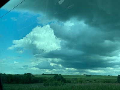 The image depicts a wide-angle view of a vast, open landscape with a dramatic sky. The lower portion of the image shows a green, grassy field with a line of dark green trees forming a dense border across the middle ground. Beyond the trees, rolling green hills are visible, suggesting farmland or pasture. The sky dominates the upper two-thirds of the frame. On the left, a patch of bright blue sky is visible, contrasting with a large, puffy, white cloud. The majority of the sky on the right is filled with dark, ominous, grey-blue clouds that appear heavy and foreboding, possibly indicating an approaching storm. The overall impression is one of a rural, natural environment with a striking meteorological phenomenon. The image appears to have been taken from a moving vehicle, as indicated by a slight reflection and a diagonal line across the top left corner, consistent with a window. There are no people or animals visible in the scene. The location is given as the city of Medyana, Russia.