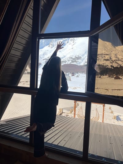 The image depicts a strong silhouette of an individual standing indoors, looking out a large window onto a snow-covered mountainous landscape in Virak, Montenegro. The individual, seen from behind, has long, straight, light-colored hair and is wearing dark loungewear, with a bare left foot partially visible. Their right arm is extended upwards, hand pressed against the window pane with fingers splayed, while their left arm rests on the window's lower frame.

The interior framing consists of dark, angled window sashes and dark corrugated paneling. Directly outside the window, a dark corrugated metal roof, partially covered in snow and scattered debris, is visible. Beyond this roof, a snow-covered outdoor area features a wooden structure suspending two distinct wicker hanging chairs. The broader exterior view encompasses a snow-covered valley with multiple small buildings and dark evergreen trees, all set against a backdrop of majestic, snow-capped mountains under a clear blue sky. The bright daylight from outside strongly backlights the interior scene, creating the silhouette.