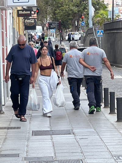 The image shows a street scene in Las Palmas De Gran Canaria, Spain, with several people walking on a sidewalk. In the foreground, an older man wearing a navy blue shirt, dark pants, and sandals walks toward the camera carrying a white plastic bag. Behind him, a young woman in a burgundy sports bra and white pants also carries a white plastic bag. Further back, a group of people is walking in the same direction. In the top of the image, two men wearing grey "ROVEL" branded t-shirts and dark work pants are walking away from the camera. Street signs and advertisements for "Mahou" and "Coca-Cola" are visible on the left side of the frame. A palm tree and various buildings are in the background. The street appears to be paved with grey tiles.