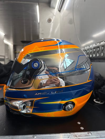 The image shows a blue, orange, and silver racing helmet with the name "ARIEL LEVI" on it. The helmet is sitting on a black surface with a white metal surface in the background. The helmet is resting on its side, and the visor is closed. The reflection of a person wearing a black shirt, a silver watch, and a black helmet is visible in the visor. The helmet is in a garage or similar location. The location is most likely the Circuit de Barcelona-Catalunya, which is located in the municipality of Montmeló, Barcelona, Spain. The coordinates provided are within the bounds of the Circuit de Barcelona-Catalunya. The helmet is likely being prepared for use by Ariel Levi, a racing driver.
