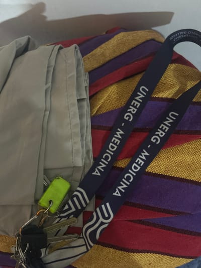 The image shows a close-up of several items. Two dark blue lanyards with white lettering reading "UNERG - MEDICINA" are prominent. The lanyards appear to be hanging from a striped textile of gold, purple, and red. Below the lanyards and the textile is a folded, light tan garment. A set of keys with a neon green tag is draped on the garment. The keys are various shapes and sizes. The image was likely taken indoors. There is no indication of the location in the city of San Juan de Los Morros, Venezuela.