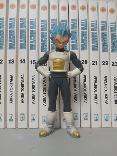 A male figure, resembling the character Vegeta from Dragon Ball, stands centered in the foreground. The figure is wearing a white and gold chest plate with gray sleeves and pants, and white boots with gold accents. Behind the figure, a row of vertically oriented books or manga volumes are arranged. The spines of these volumes are predominantly white with the text "DRAGON BALL" and "AKIRA TORIYAMA" in a blue font. Numbers such as "13", "14", "15", "16", "20", "21", "22", and "23" are visible on some of the spines. The bottom portion of the book spines show logos with the text "EDIZIONI STAR COMICS". The background appears to be a plain, light-colored surface. The overall scene is indoors, likely on a shelf or table. The image composition suggests a product display or collection.