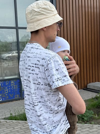 A man wearing a bucket hat and a printed shirt is holding a baby with a pacifier, in Речицы, Russia. The location is indicated by the blue pattern on the wall and the architecture in the background. The man is looking at the baby. The image is taken from a medium distance, with the subjects filling most of the frame. A building with brown horizontal siding is partially visible behind the man and child. The baby is wearing a white hat.