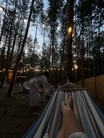In Ivachiv Horishnii, Ukraine, a serene campsite is illuminated by fairy lights strung between tall trees. A person is relaxing in a striped hammock, their legs extended, while another individual in a white robe stands near a campfire, tending to the flames. In the background, a wooden structure resembling a sauna or cabin is visible. The scene is set in a wooded area, with pine trees forming a dense canopy overhead. The twilight sky is a soft blue, indicating the transition from day to night.