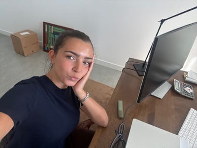 A woman with dark hair pulled back, wearing a dark blue short-sleeved top and a silver watch, is seated at a desk. She is looking directly at the camera with her hand resting on her face. The desk holds a white keyboard, a white Apple laptop, a gray calculator, a white mouse, a green highlighter, and a large computer monitor on a stand. In the background, there's a framed abstract artwork and a cardboard box with "H&M" printed on it. The location is not discernible from the image. It is not possible to determine if the location is in the city of Torres Vedras, Portugal.