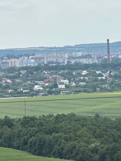 A distant cityscape is visible under an overcast sky, featuring a variety of buildings ranging from multi-story apartment complexes to smaller houses with red and gray roofs. A tall, reddish-brown industrial chimney stands prominently on the right side of the image, towards the middle distance. In the foreground, rolling green fields are separated by dark green tree lines. Power lines are visible in the lower part of the image. The overall scene suggests a rural or semi-rural environment on the outskirts of a city. The location is identified as Kolubaivtsi, Ukraine.