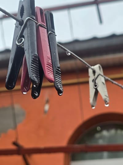 This is a close-up, outdoor, eye-level shot of several wet clothespins hanging on a wire against a blurry orange wall and a dull sky. Three black and one pink clothespin are clustered together on the left, with water droplets hanging from their tips. A single light-colored clothespin is further down the wire on the right, also with a water droplet. The scene is likely in Nova Kakhovka, Ukraine, given the context of the request. The focus is sharp on the clothespins, with the background out of focus. The image conveys a sense of dampness and overcast weather.