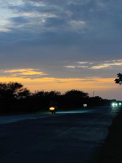 The image depicts a dusky scene on a road, likely in or near Dattapur, India. The sky is a gradient of deep blue at the top, transitioning to hues of orange and yellow closer to the horizon, indicating either sunrise or sunset. Wispy clouds are scattered across the sky. Silhouetted against the lighter sky are dark trees lining the sides of the road.
In the foreground, the asphalt road stretches into the distance, appearing dark and somewhat worn. On the road, a motorcycle with its headlights illuminated is visible, moving away from the viewer. Further down the road, the headlights of another vehicle or vehicles are also visible, creating bright points of light against the dim environment. The presence of vehicles on the road suggests this is a traveled route. The overall atmosphere is one of quietude and the transition between day and night.