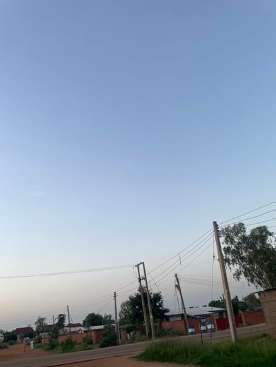 The image depicts a view of a street in Wauro Jabbe, Nigeria, under a clear, pale blue sky that transitions to a lighter hue near the horizon. In the foreground, a dirt road curves to the right, bordered by green grass and some bushes. Several utility poles with overhead wires crisscross the scene, some appearing weathered. Beyond the road, a collection of brick buildings with red roofs are visible, interspersed with trees and vegetation. Two individuals, dressed in light-colored traditional clothing, are walking away from the viewer on the dirt road. In the mid-ground, a street sign with blue and white stripes reads "SUNTAI RD." The overall impression is that of a quiet, semi-urban street in a Nigerian town.
