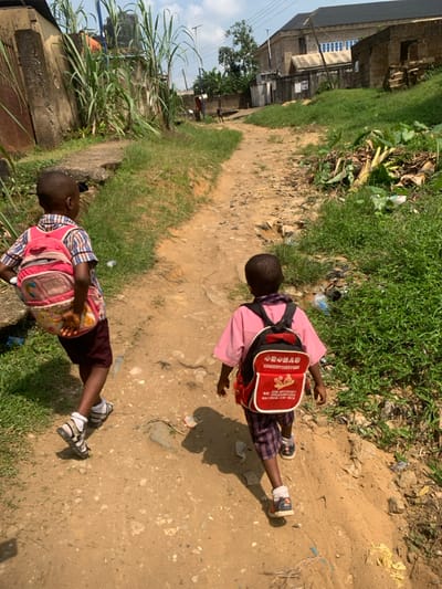 Two young children are observed from behind, walking away from the camera on a sunlit, unpaved dirt path in Anantigha, Nigeria. The path is uneven, composed of light brown soil, small rocks, and debris.
The child on the left wears a pink and white plaid shirt, dark shorts, white socks, and white sandals, carrying a faded pink backpack. The child on the right wears a pink shirt, plaid shorts, white socks, and dark sandals, carrying a red backpack with white text, including what appears to be Chinese characters and phone numbers. Both children have short, dark hair.
To the left of the path, a concrete structure, possibly a drainage culvert or retaining wall, is visible, partially obscured by tall green plants resembling reeds or sugarcane. Further left, a corrugated metal fence and parts of a concrete building are present. To the right of the path, a grassy embankment slopes upward, containing green foliage and scattered litter, including plastic items and plant waste.
In the mid-ground and background, the dirt path continues, bordered by various structures. On the right, a multi-story building with a dark roof and lighter-colored walls is prominent, alongside other less defined buildings and fences. Utility poles with overhead wires are visible against a bright, partly cloudy sky. Distinct shadows of the children are cast on the path by the strong sunlight, indicating a clear day. Another distant figure is barely discernible further down the path.