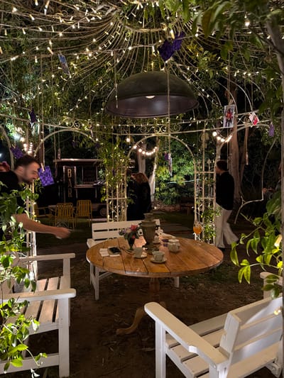 This image depicts an outdoor evening scene, likely a garden or patio event, illuminated by numerous string lights. The setting is adorned with white ornate metal archways or pergolas, draped with green foliage. Small photographs are clipped to the string lights, adding a personal touch.
In the foreground, a round wooden table is set with a traditional samovar, white teacups, a teapot, and a small floral arrangement. A smartphone and other items are also visible on the table. Two white wooden slatted chairs are positioned around it.
Several people are present. A man with a beard, wearing a dark shirt, is prominent in the foreground, looking upwards and reaching his arm out. Another person with reddish-brown hair is partially visible behind the table. Further back, a person in a black top and white pants stands near one of the illuminated archways.
In the background, a structure with the word "BAR" prominently displayed is visible, along with several yellow metal chairs. The atmosphere is warm and inviting, created by the abundant string lights. The scene suggests a social gathering or event, possibly involving a tea service or drinks. The time of day is night, with artificial lights providing the primary illumination. The location is Nir Eliyahu, Israel.