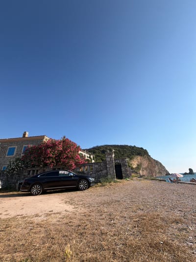A black four-door sedan is parked on a gravel lot with dry grass in front of a stone house and a stone archway. The house has blue shutters and a red-tiled roof. A large bush with pink flowers is in front of the house. To the right, a rocky hill slopes down to the sea. The sea is blue and calm, with a few people and an umbrella on the shore. The sky is clear and blue.