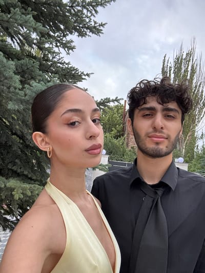 The image displays two individuals, one female and one male, posing outdoors in what is identified as the city of Nor Hachin, Armenia. The female, positioned on the left and slightly closer to the foreground, has dark hair pulled into a slicked-back bun. She wears a light yellow, V-neck, halter-neck garment and a small gold-toned hoop earring in her visible right ear. Her head is angled slightly to her left, with a neutral facial expression. The male, positioned to the right and slightly behind the female, has dark, curly hair and light facial hair. He is attired in a black collared shirt with a loose black tie. His gaze is directed towards the camera, maintaining a neutral expression. The immediate background consists of dense green foliage, including evergreen trees on the left and a deciduous tree with bare branches on the right. Spherical white outdoor light fixtures are visible on pillars behind the individuals, and a patterned paved surface is present in the lower left. The sky overhead is overcast.