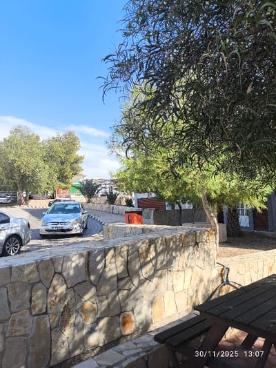 The image depicts an outdoor scene in Gönyeli, Northern Cyprus / Cyprus, captured on November 30, 2025, at 13:07. In the immediate foreground, a dark wooden picnic table with benches is partially visible in the lower right. A light-colored stone retaining wall, constructed with irregularly shaped blocks, runs from the center foreground towards the left and then turns right, defining the edge of a paved area.
Behind this wall, a paved driveway or parking area curves to the left. A silver Peugeot sedan, identifiable by its front grille and badging, with license plate "CK 735," is parked facing the viewer on this paved surface. To the far left, the rear section of another silver sedan is partially visible. Further along the paved area and behind the stone wall, an orange rectangular container is positioned.
The environment features various trees with green foliage, including a prominent tree with long, narrow leaves on the right side, partially obscuring the view. Additional trees and shrubs are scattered throughout the mid-ground and background. In the background, several low-rise buildings or structures are visible, including one on the right with a reddish-brown section and white window or door frames. The sky is clear blue with scattered white clouds, and distant, low-lying terrain is discernible on the horizon. No individuals or animals are present.