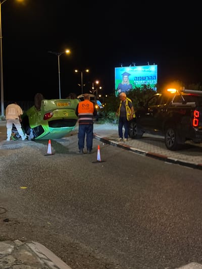 At night in Kiryat Motzkin, Israel, a green car lies overturned on the side of a road, surrounded by orange cones. Two men in reflective vests stand nearby: one is inspecting the overturned car, and the other is standing near a black tow truck. A billboard with an advertisement in Hebrew is visible in the background. A person in white clothing is near the car's wreckage. Streetlights illuminate the scene. The road is paved and bordered by a curb.