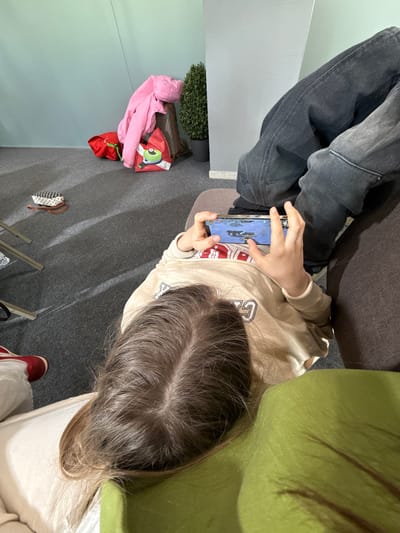 A person with long brown hair is sitting and playing a game on a smartphone. The person is wearing a beige sweatshirt with white lettering. They are seated between a gray and a green sofa. Another person's legs are visible in gray jeans. A pink jacket, a red bag, and a small black and white purse are on the gray carpet. A potted plant is nearby. A red shoe is also visible. The setting may be indoors based on the lighting and the presence of furniture, but the exact location within Peresvet, Russia, cannot be determined from the image.