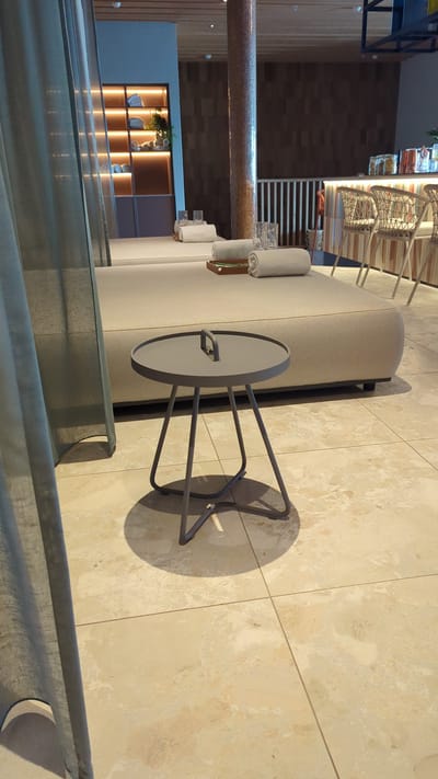 The image depicts an indoor spa or relaxation area. In the foreground, a small, round, dark gray side table with three slender legs stands on a tiled floor. To the left, a portion of a light gray curtain is visible. Behind the table, a large, light beige chaise lounge is positioned, with two rolled towels and two glasses placed on it. The lounge appears to be part of a seating area. In the background, a shelving unit illuminated with warm lights displays folded towels and some greenery. Further back, a bar area with chairs and a counter is visible, with decorative wooden panels on the wall behind it. The overall ambiance suggests a tranquil and luxurious setting. The specific location in Larkollen, Norway is not discernible from the image itself.